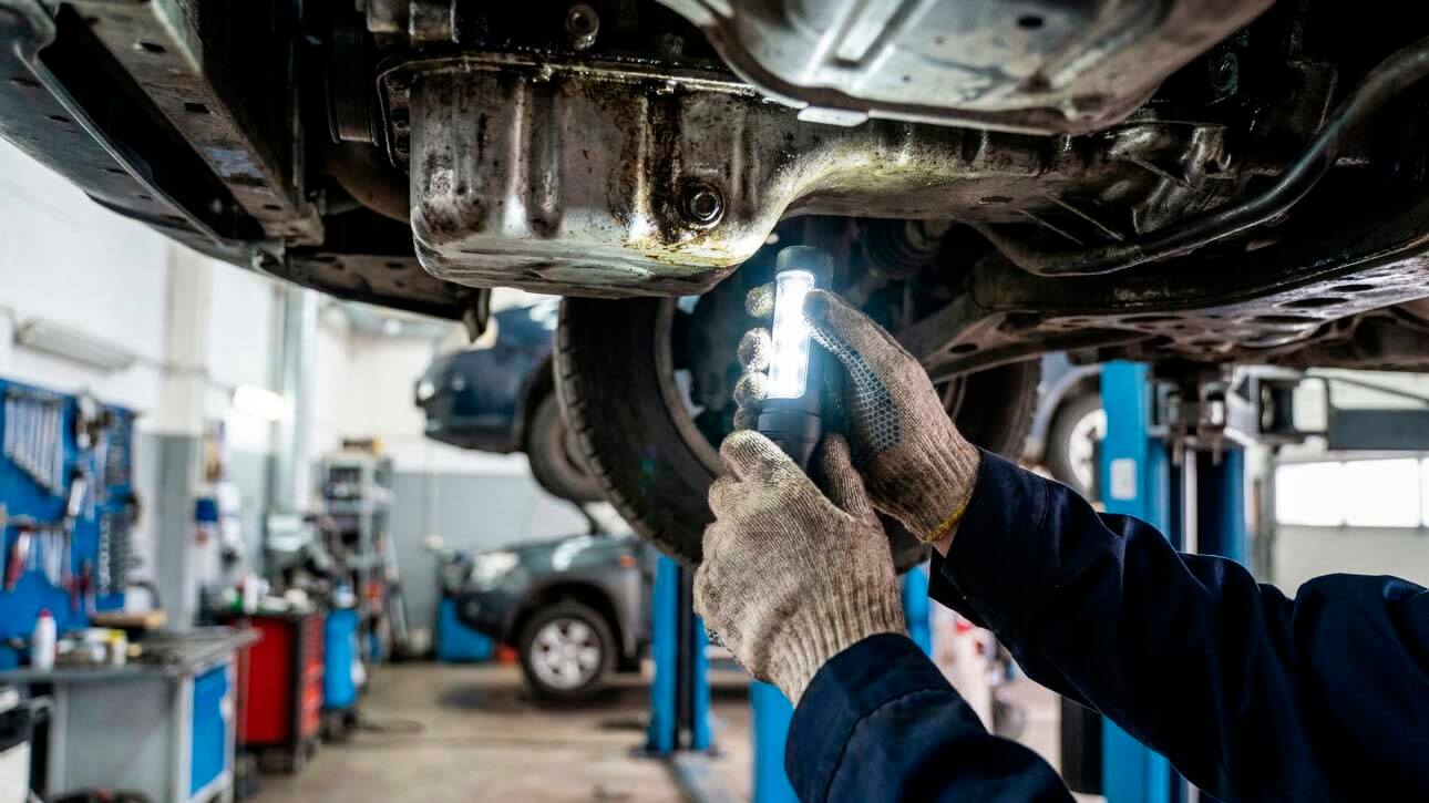 автомеханик светит фонариком на двигатель снизу автомобиля